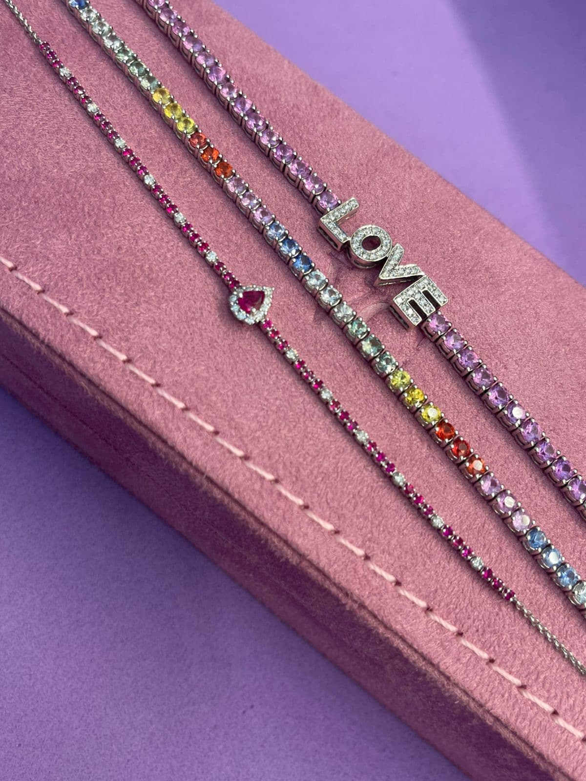 Pulseiras riviera de safiras multicoloridas e pingente LOVE em diamantes, sobre camurça rosa e fundo lilás